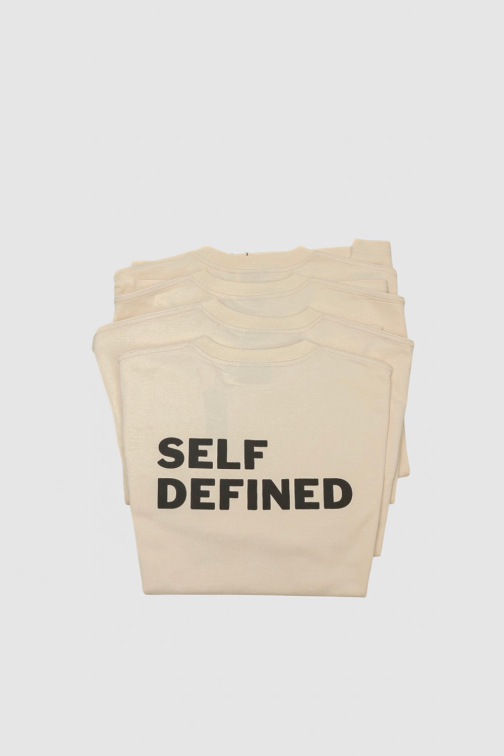 Stack of folded seedpearl DROP 001 tees with SELF DEFINED on top