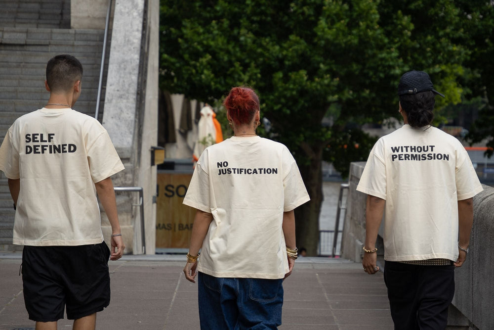 Three models on the South Bank, London, wearing DROP 001 tees — SELF DEFINED, NO JUSTIFICATION and WITHOUT PERMISSION raised puff back prints